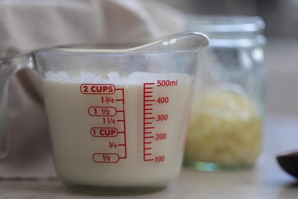 2 cups of whole milk in a measuring glass with a jar of kefir grains in the background