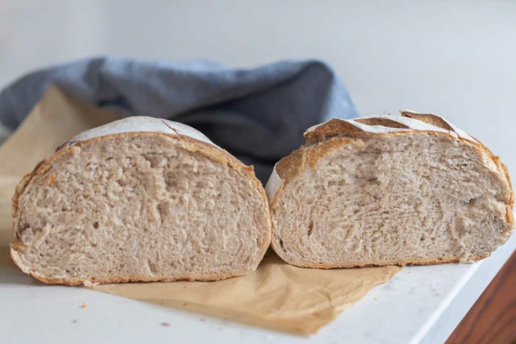 A loaf of low hydration sourdough bread cut in half on a piece of parchment paper with a blue towel in background