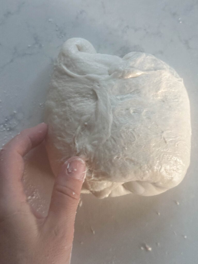 A woman's hand shaping a small batch sourdough bread dough ball on a white countertop that has been floured