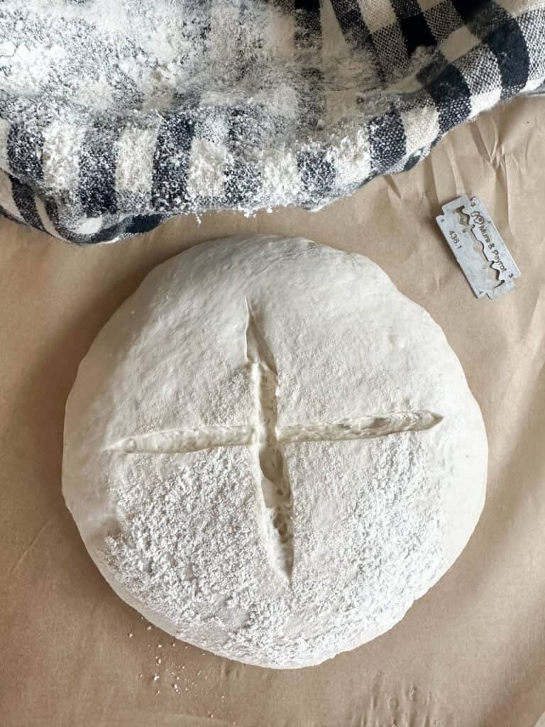 A small batch sourdough bread dough ball on a piece of parchment paper next to a blue and white checkered tea towel, scored and ready to bake