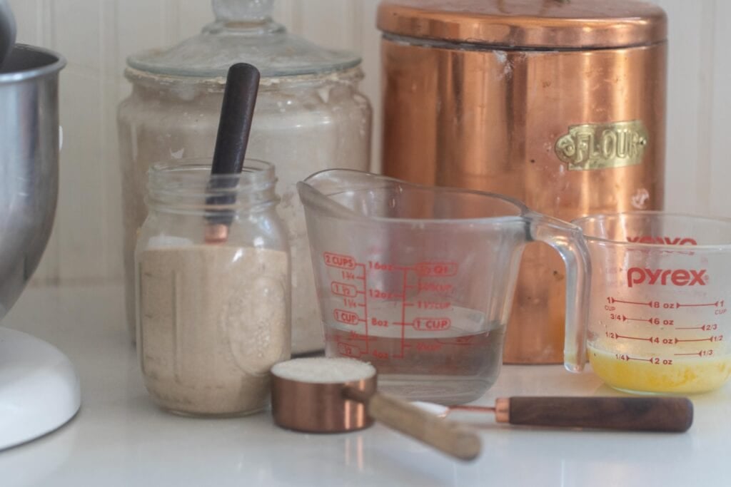jars and containers full of ingredients to make sourdough discard rolls like flour, water, Sourdough starter, yeasts, and butter.