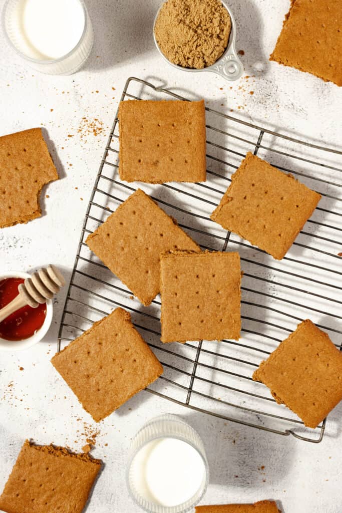 9 sourdough graham crackers spread over a wire cooling wrack on a white countertop surrounded by glasses of milk, brown sugar, and honey