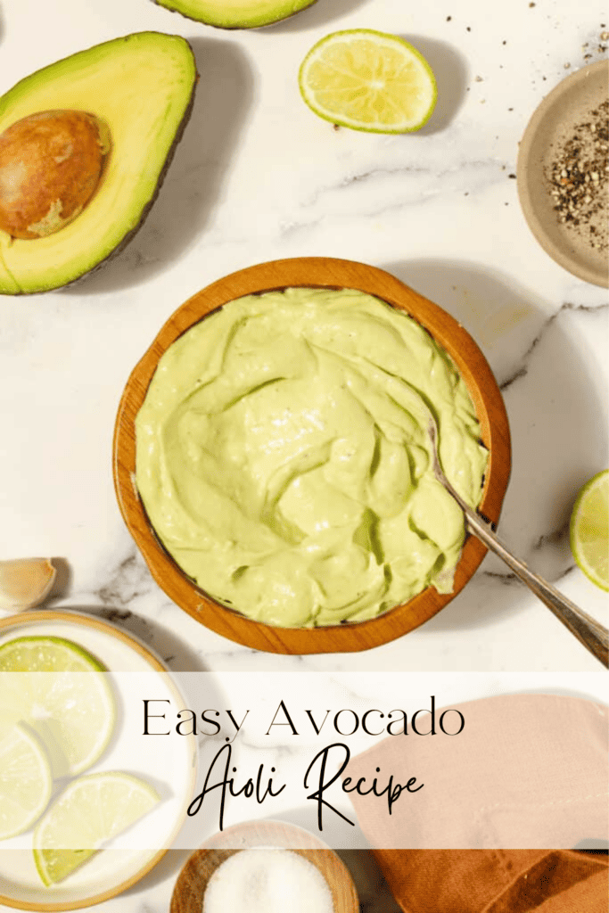 Avocado aioli in a wooden bowl with a silver spoon on a white countertop, surrounded by avocados and limes.