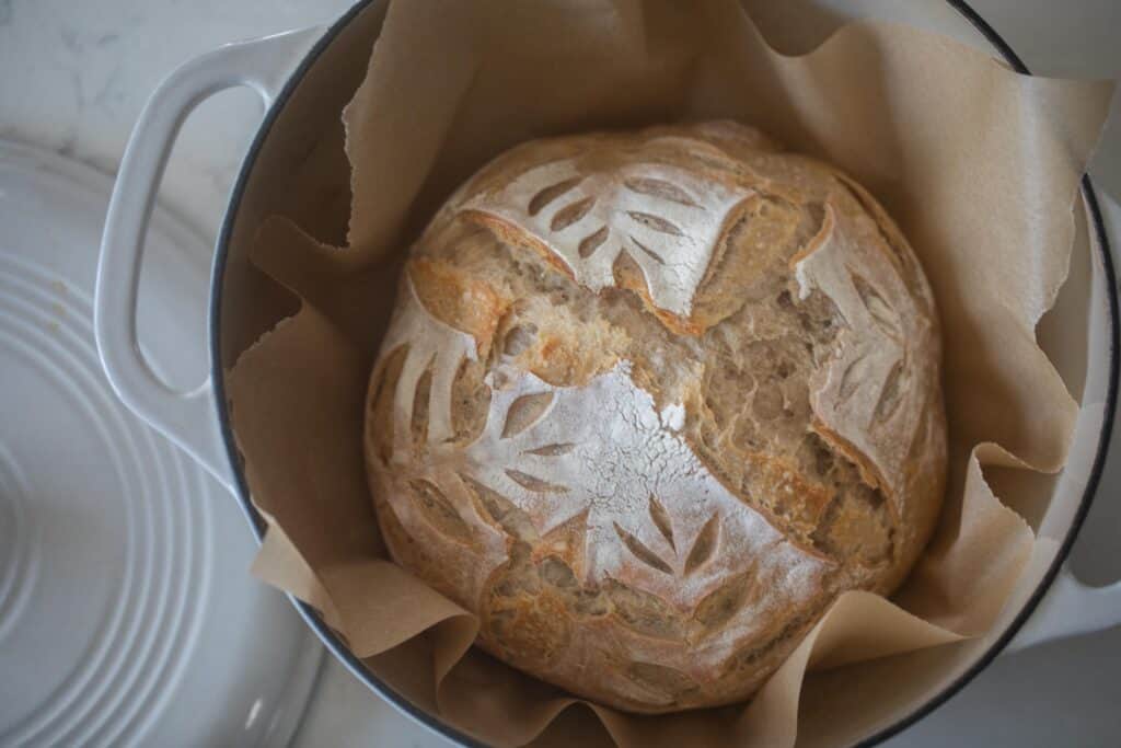 A baked sourdough loaf with a golden brown crust placed on parchment paper inside a white dutch oven with the lid to the side