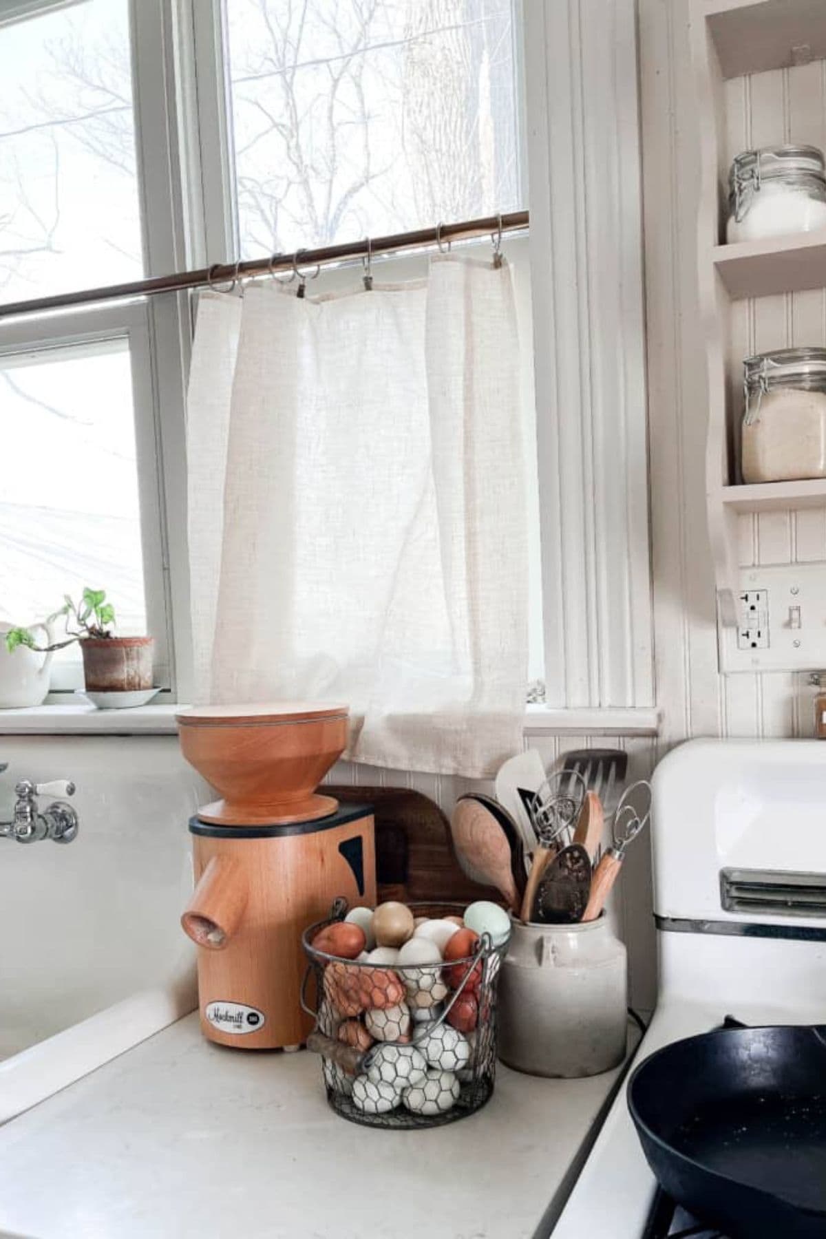 A mockmill on a kitchen counter next to a window and a bucket of eggs.