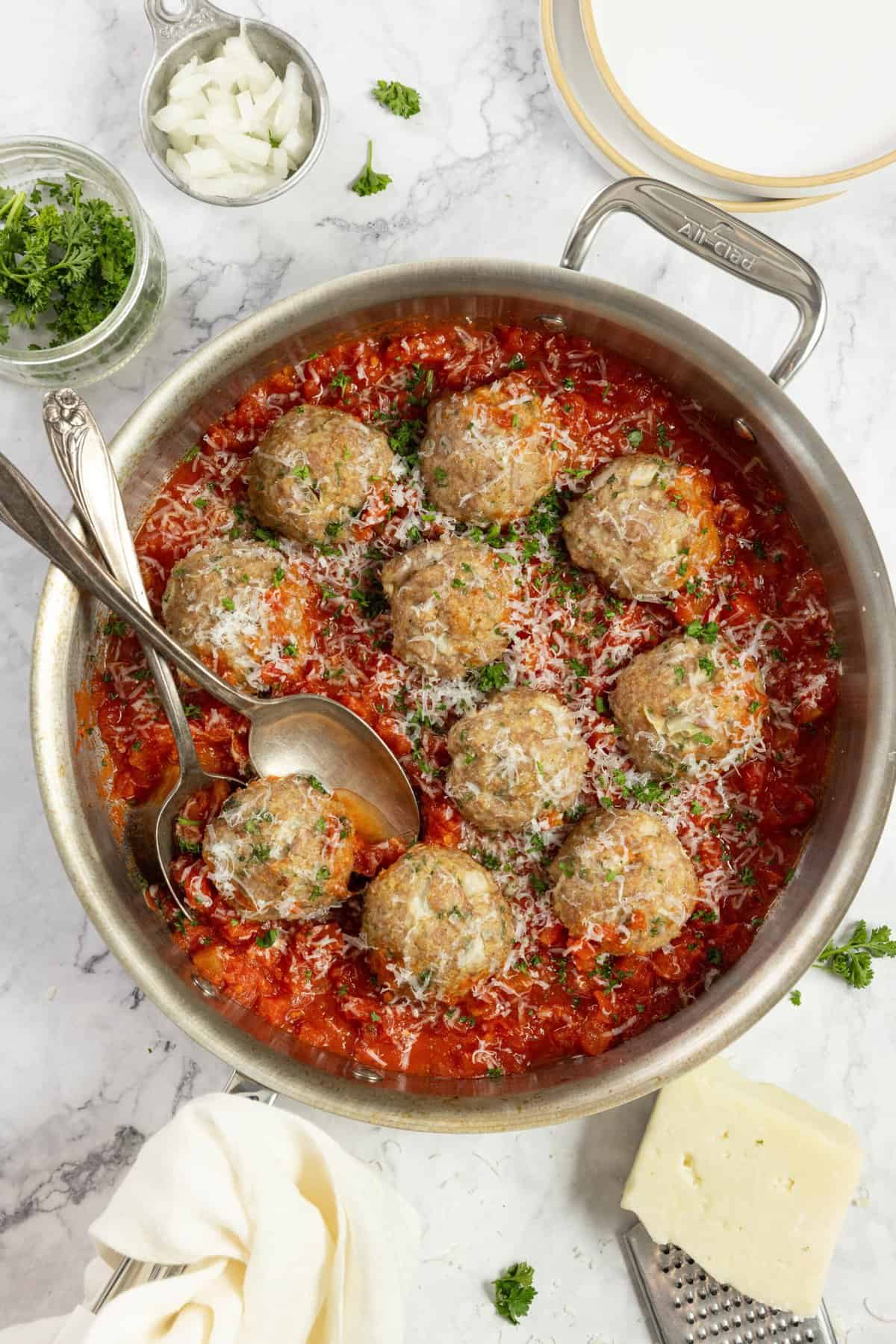 overhead photo of sausage meatballs in tomato sauce in a stainless frying pan with ingredients surrounding the pan