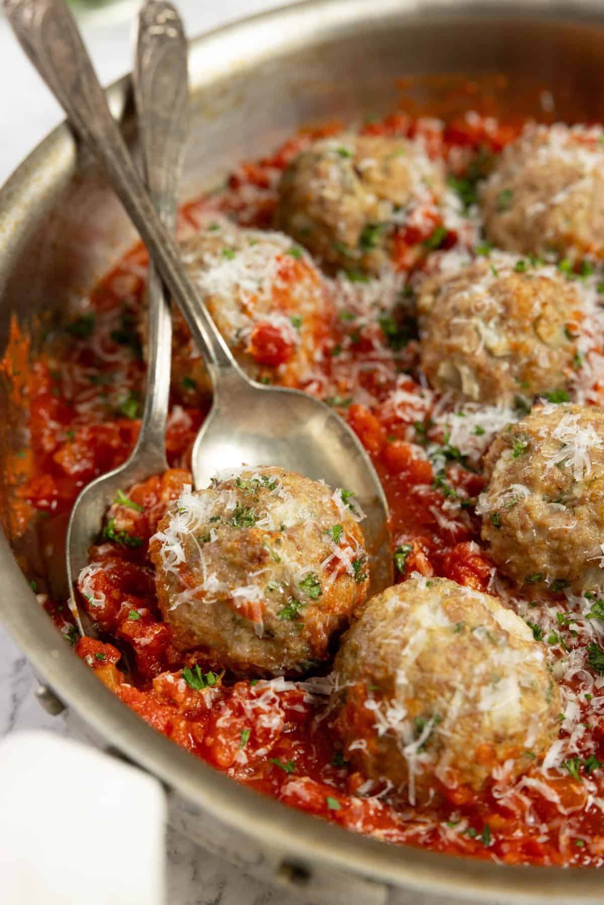 close up picture of sausage meatballs in red sauce in a stainless pan with spoons in the meatballs
