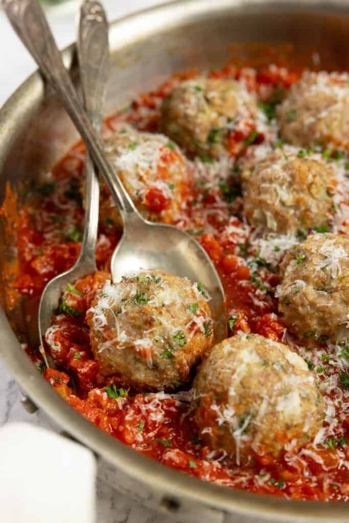 close up picture of sausage meatballs in red sauce in a stainless pan with spoons in the meatballs