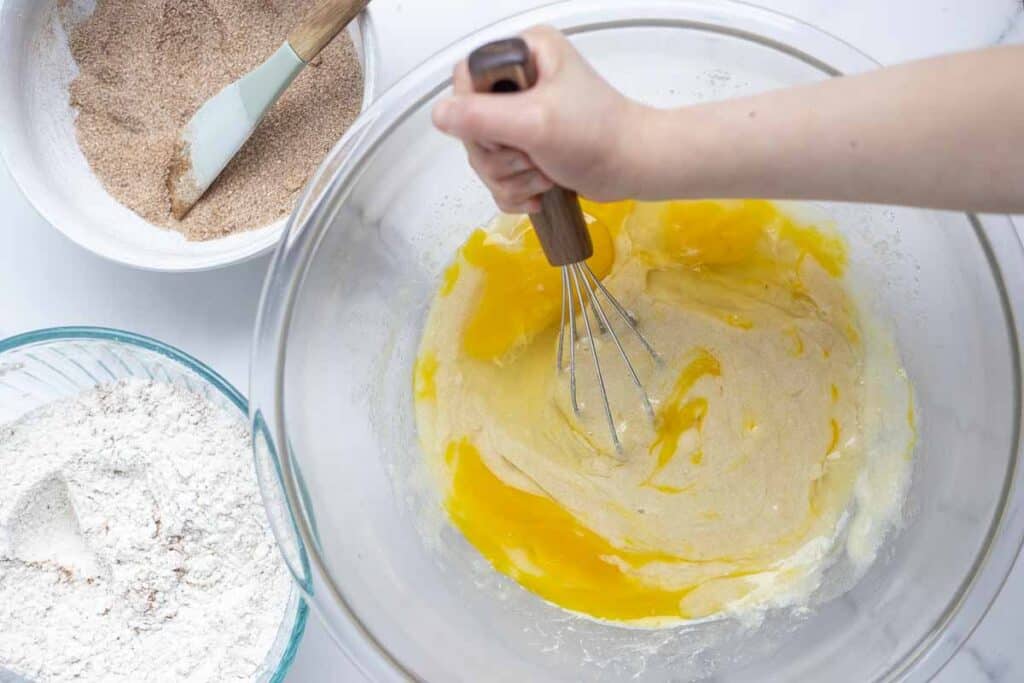 hand holding whisk and whisking together eggs, sourdough starter and butter. More bowls of ingredients surrounding