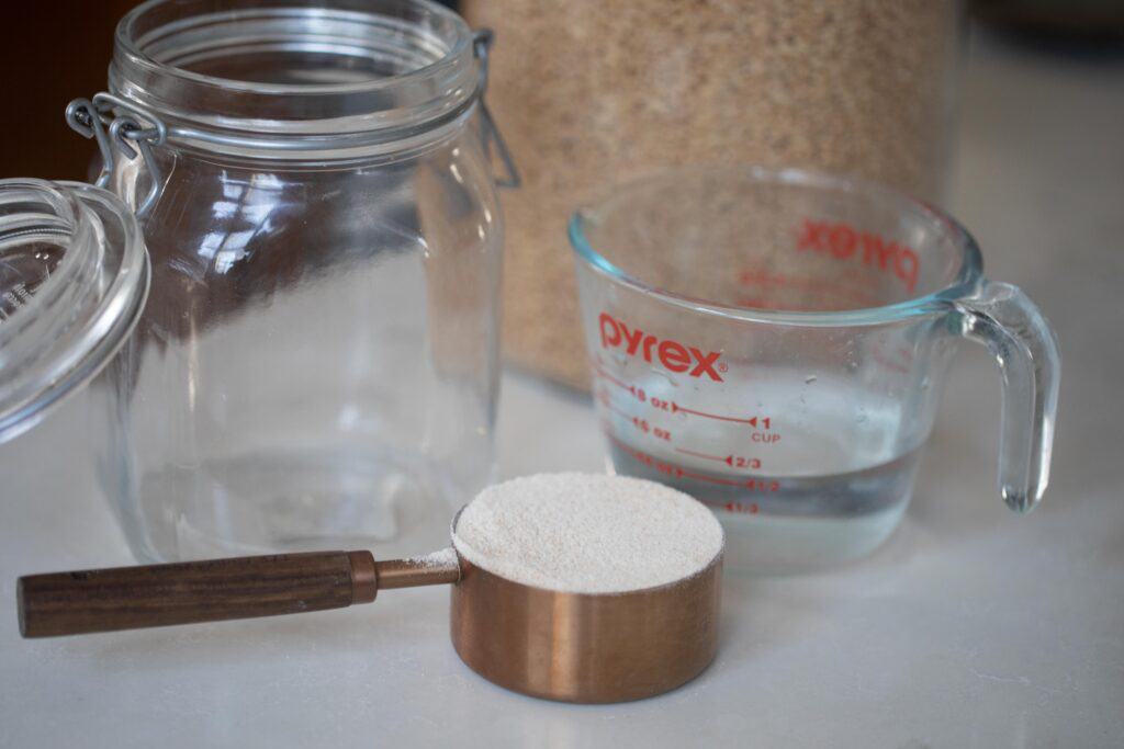 a clean flip top jar, glass pyrex measuring cup of water, and a copped and wood dry measuring cup full of freshly milled wheat flour