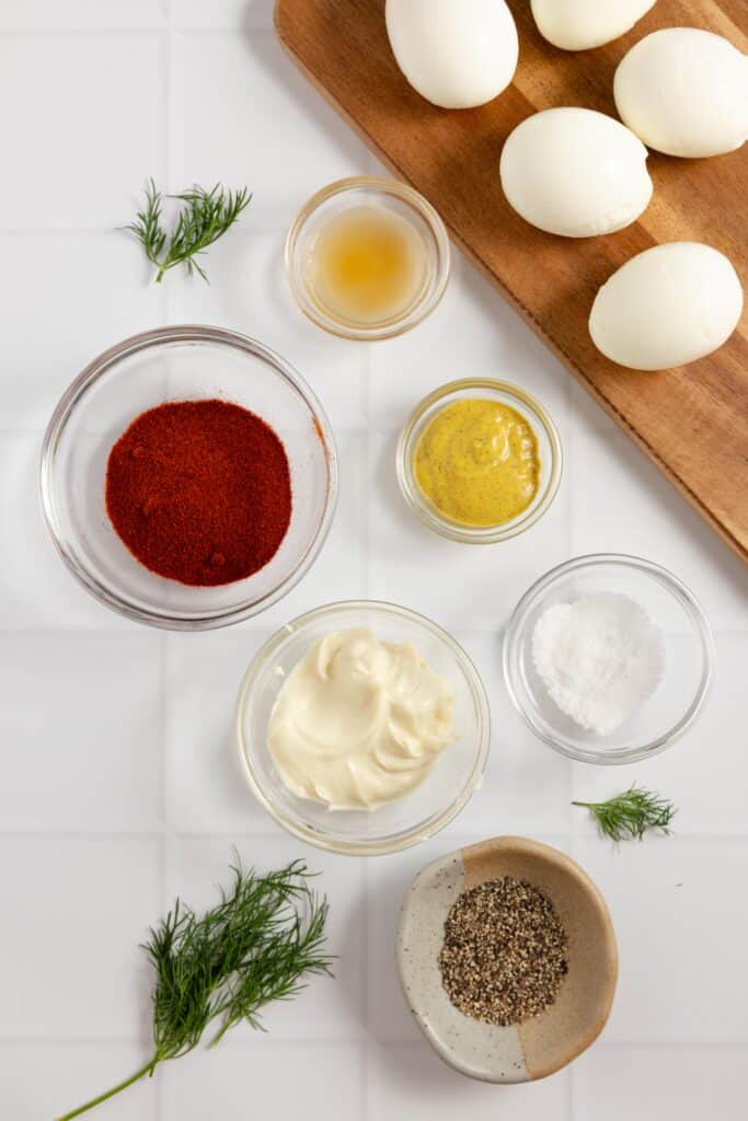little glass bowls with ingredients on a white countertop with eggs on a wooden cutting board to the right