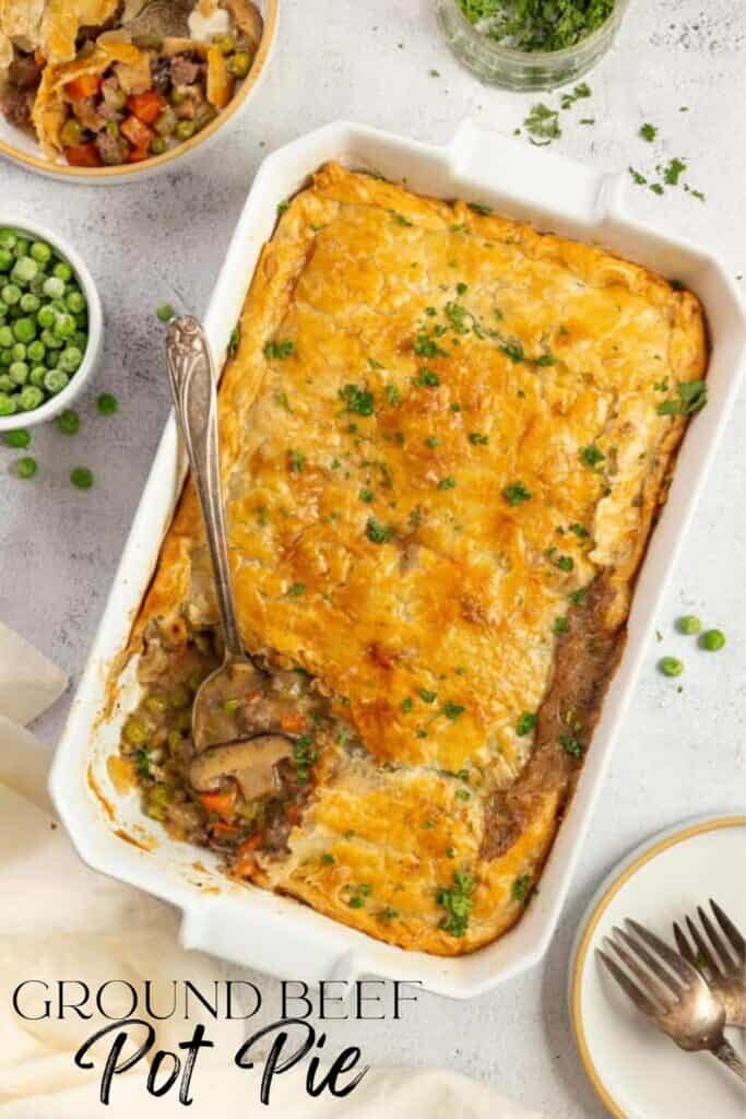 overhead photo of ground beef pot pie topped with a golden brown pie crust and parsley with a spoon in the corner of the pie. The baking dish sits on a white countertop surrounded by ingredients