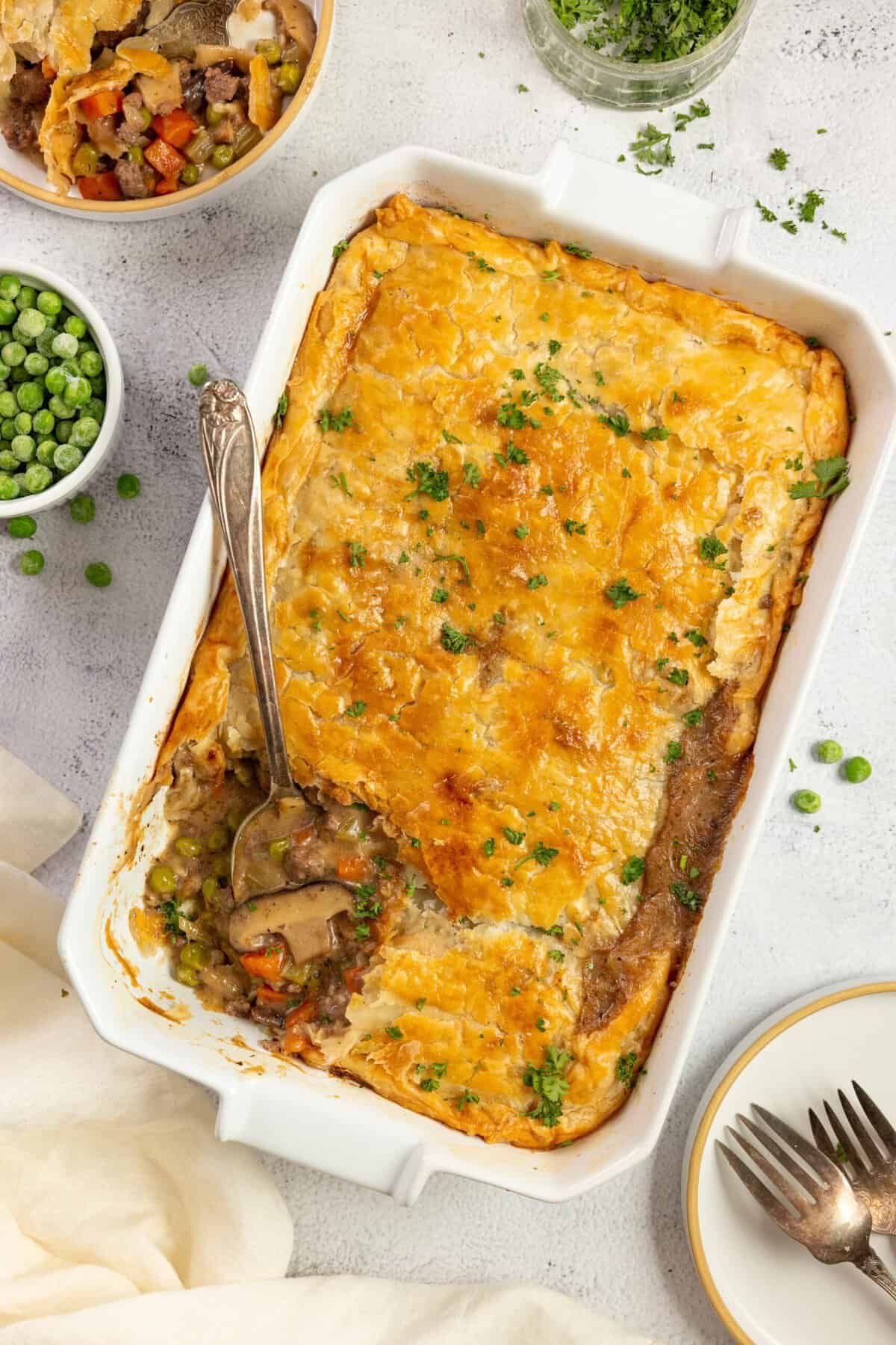 overhead photo of ground beef pot pie topped with a golden brown pie crust and parsley with a spoon in the corner of the pie. The baking dish sits on a white countertop surrounded by ingredients