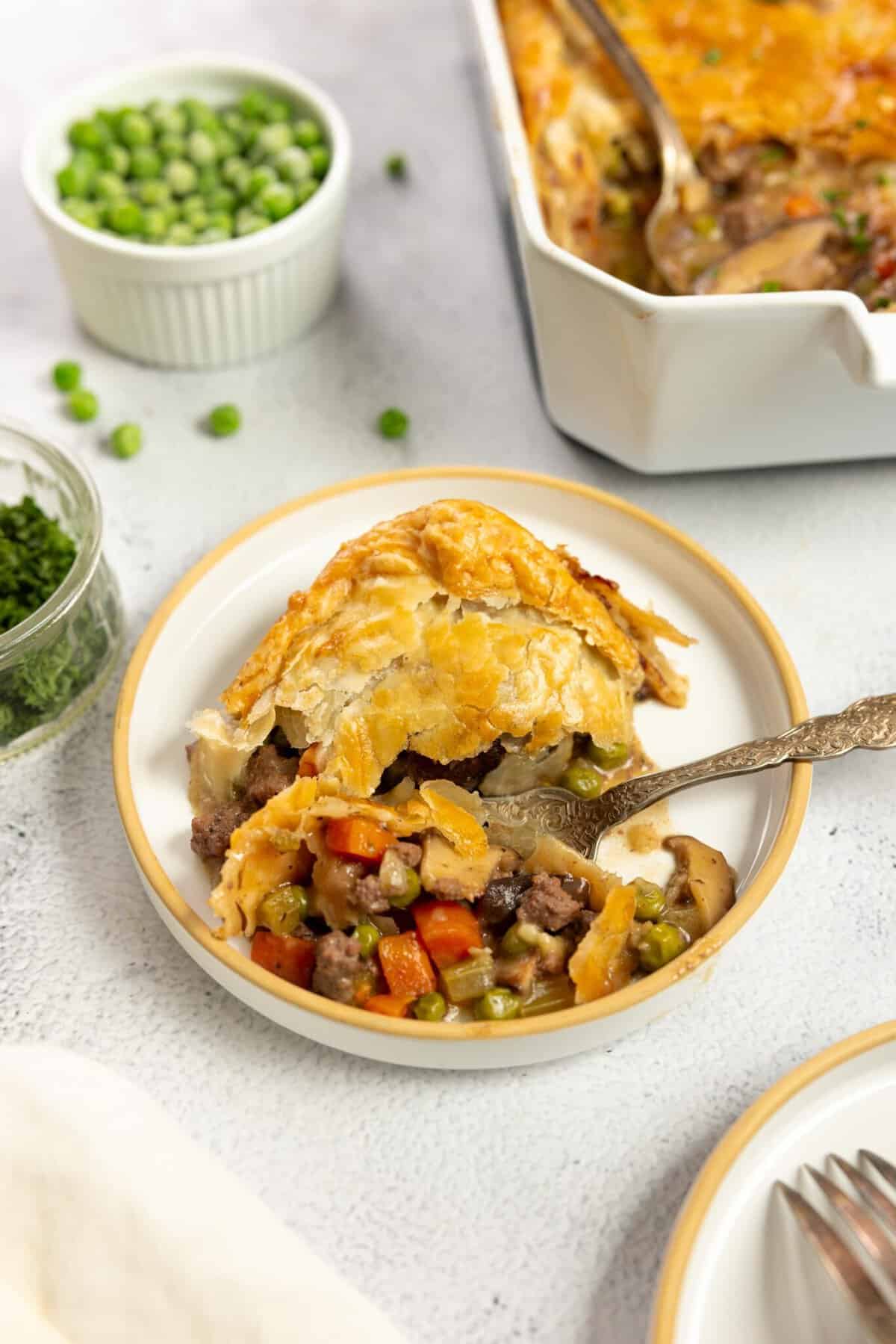 plate full of ground beef pot pie on a white plate with a baking dish of more pie and a bowl of peas in the background