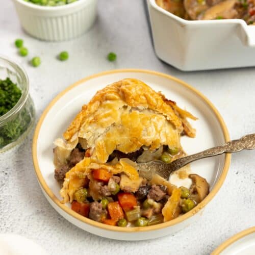 plate full of ground beef pot pie on a white plate with a baking dish of more pie and a bowl of peas in the background