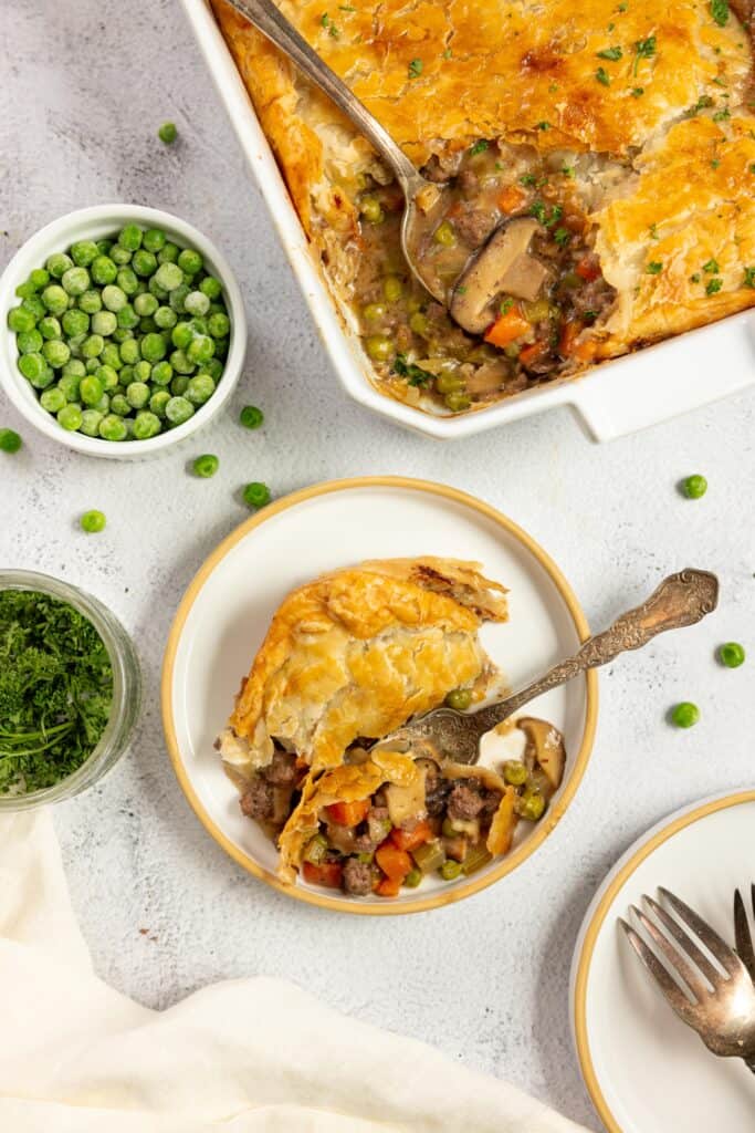 overhead photo of a plate with a spice of ground beef pot pie with a fork. A small bowl of peas and a baking dish with the remaining pie in in the background