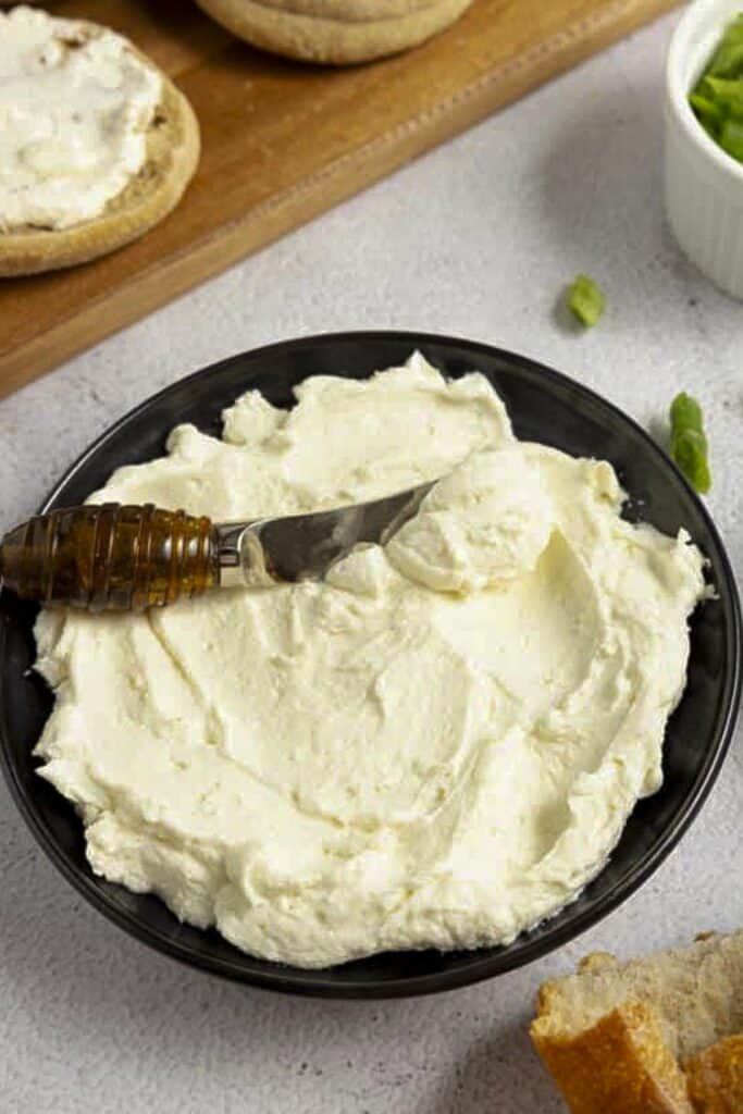 Close up picture of cream cheese in a black dish with a little knife. Bagels are in the background