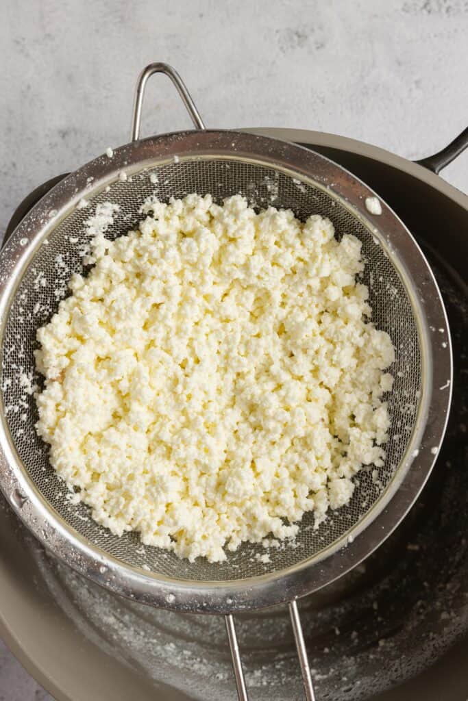 A fine mesh strainer above a bowl straining out the whey from the curds to make homemade cream cheese