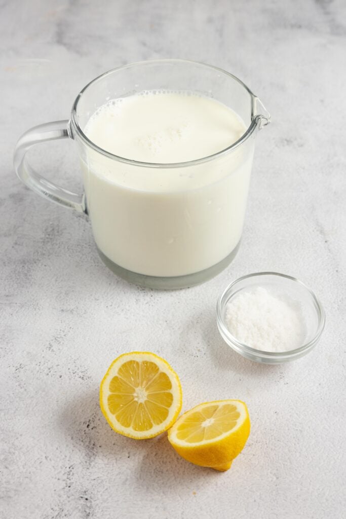 The ingredients for homemade cream cheese, including a lemon cut in half, a glass bowl with salt and a glass measuring cup with whole milk all on a white countertop