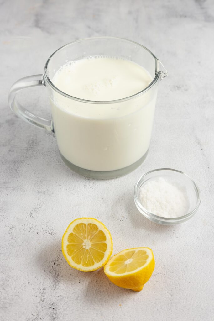 The ingredients for homemade cream cheese, including a lemon cut in half, a glass bowl with salt and a glass measuring cup with whole milk all on a white countertop