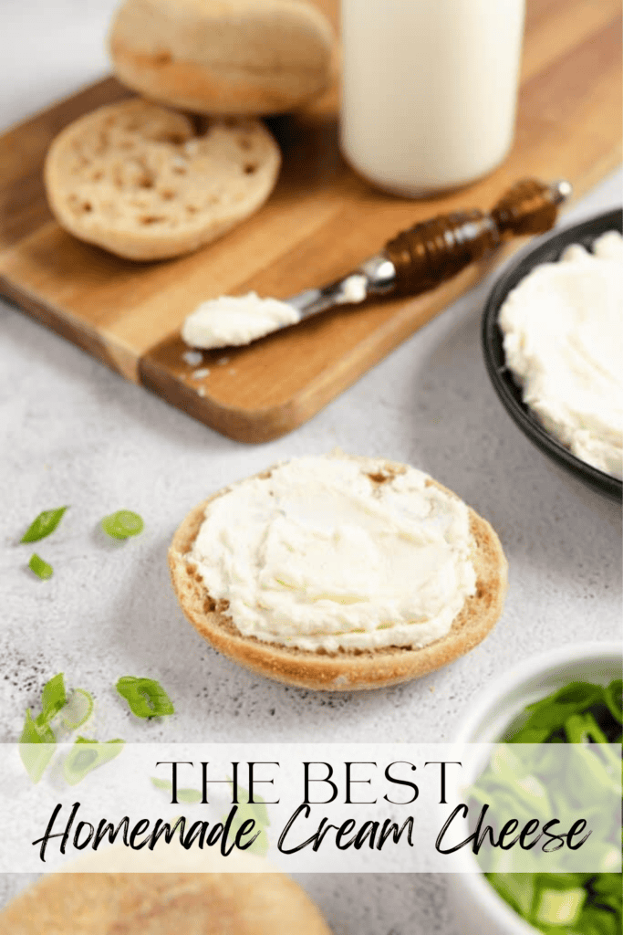 An english muffin with homemade cream cheese spread on top with a black bowl of it and a spreading knife and wooden cutting board in the background