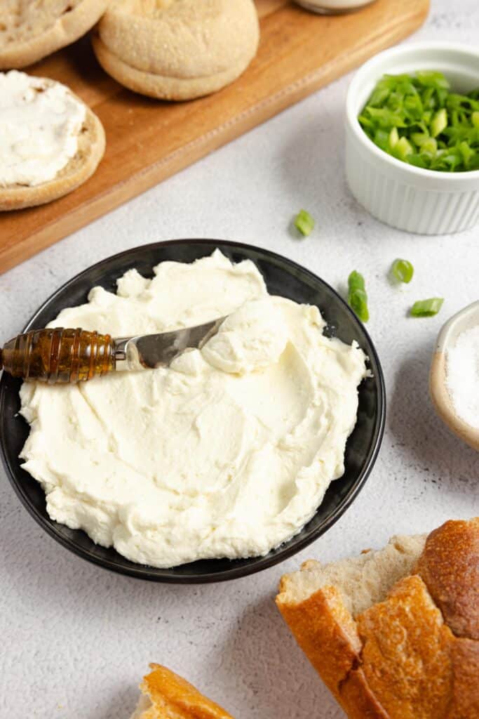 a bowl of homemade cream cheese with a spreading knife in it and other food to eat with the cream cheese surrounding it on a white countertop and wooden cutting board