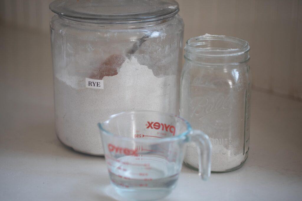 a large glass jar with flour, a emo=pty mason jar, and a liquid measuring cup of water on a white quartz countertop