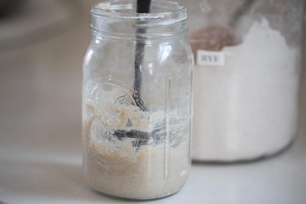 a spoon in a glass mason jar of flour and water. More flour is in the background