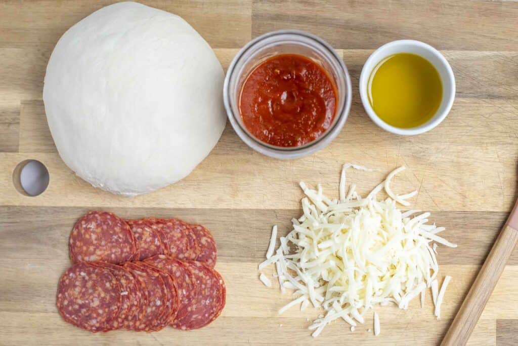 overhead photo of pizza dough, pizza sauce, olive oil, pepperoni, and cheese on a wood cutting board