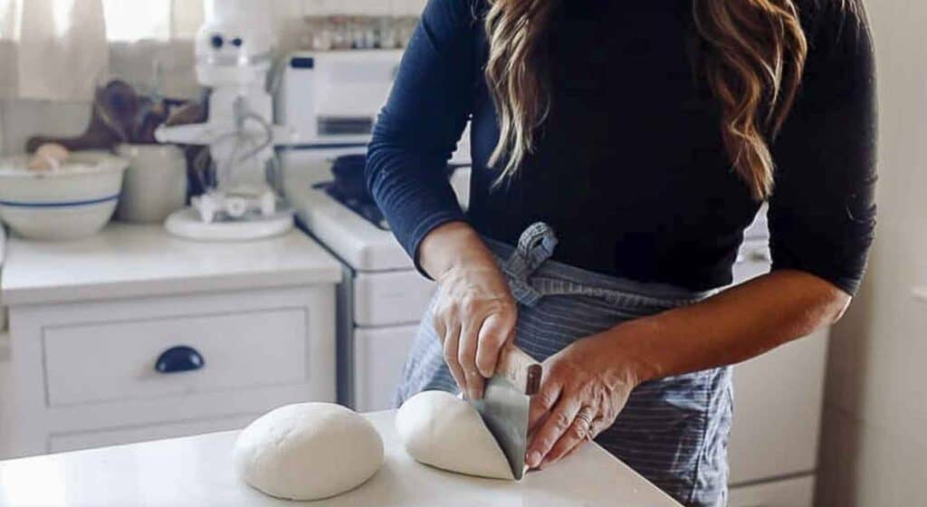 a women cutting sourdough pizza dough with a bench scraper on a quartz countertop