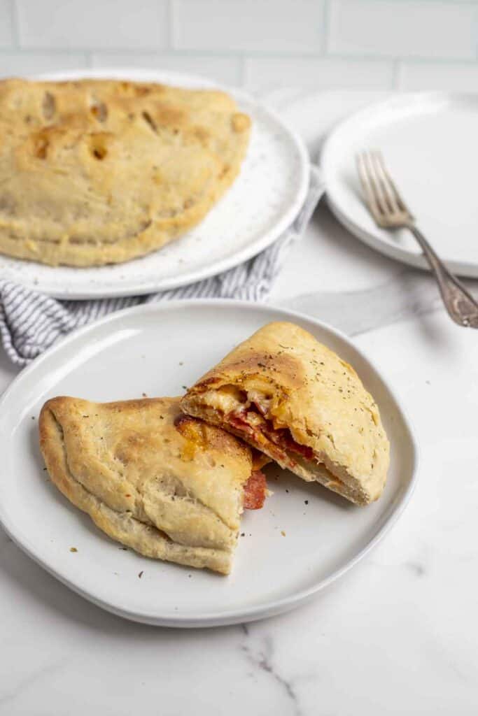 sourdough calzones cut in half on a cream colored plate on a marble countertop. More calzones are in the background