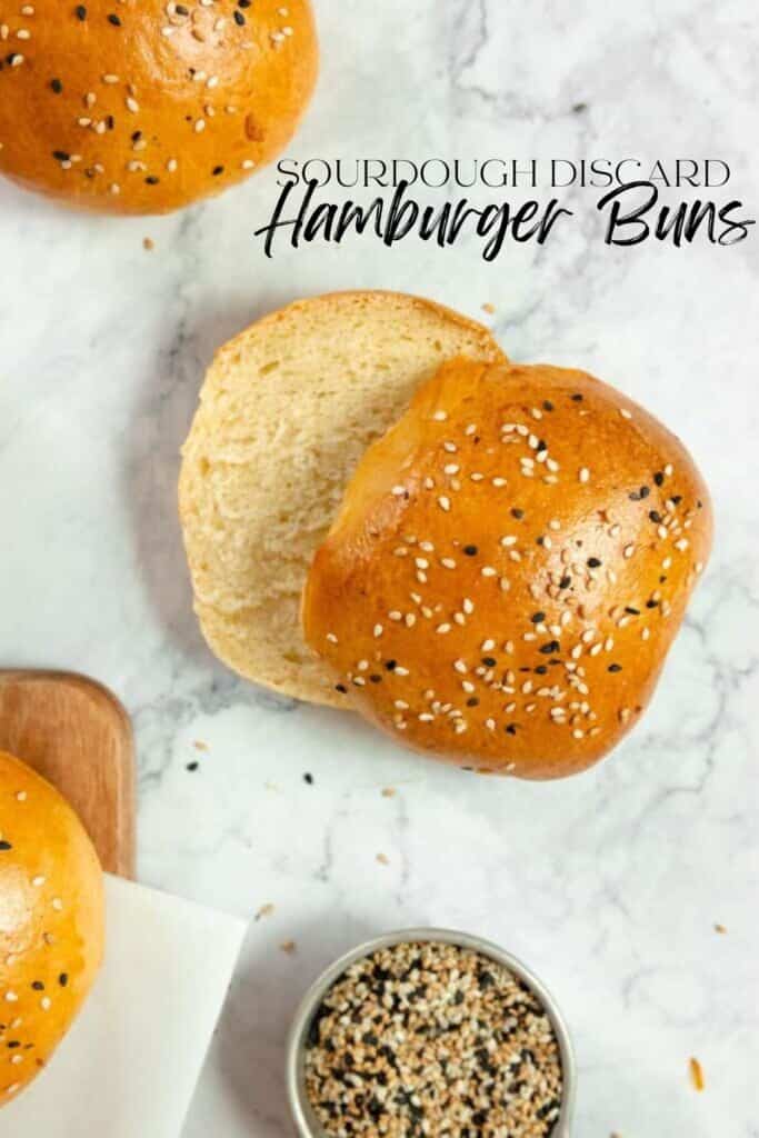 a sourdough discard hamburger bun sliced in half on a marble countertop with two more buns to the left and a jar of sesame seeds in front.