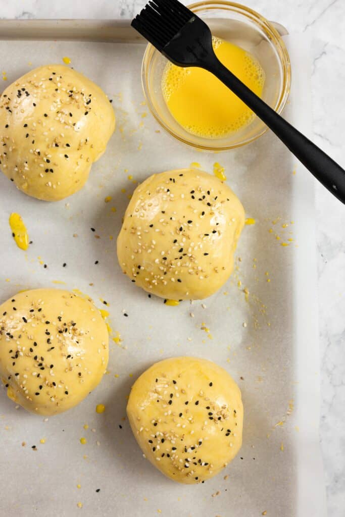 hamburger bun dough with a egg wash and topped with sesame seeds on a parchment lined baking sheet.
