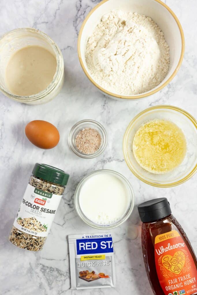 jars and bowl so flour, melted butter, sourdough starter, salt, yeast, a bottle of honey, sesame seeds and an egg on a marble countertop