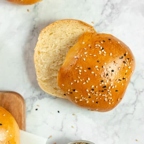 a sourdough discard hamburger bun sliced in half on a marble countertop with two more buns to the left and a jar of sesame seeds in front.