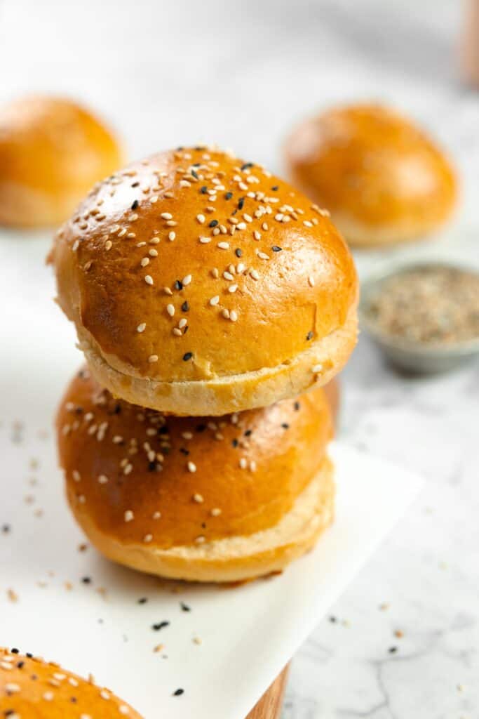 two sourdough discard buns stacked on top of each other on a marble countertop. more buns and sesame seeds are in the background