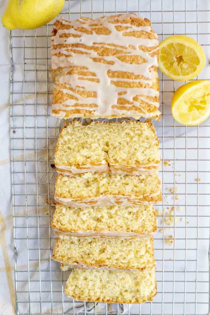 overhead photo of sourdough lemon discard bread loaf topped with icing on a wire rack. Half of the loaf sliced. lemons surround the bread