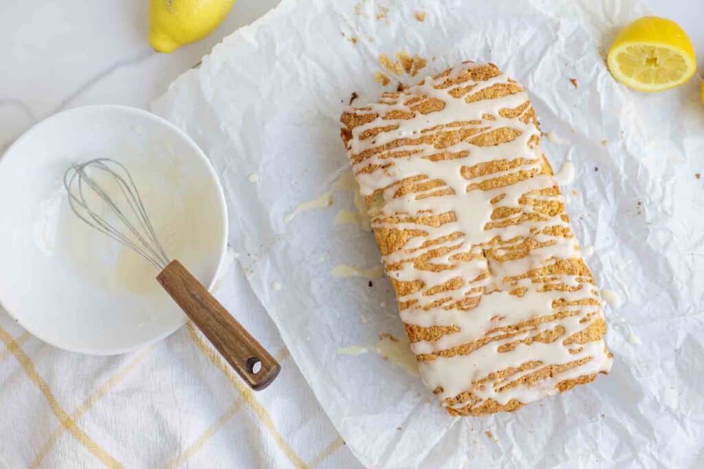 loaf of lemon bread on parchment paper with a lemon glaze sprinkled over top. A bowl of lemon glaze with a whisk is the left.