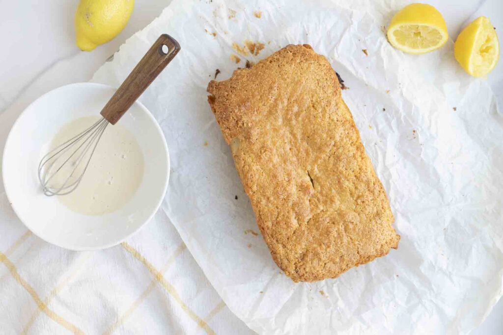 baked loaf of bread on parchment paper. A bowl of lemon glaze with a wooden spatula to the left.