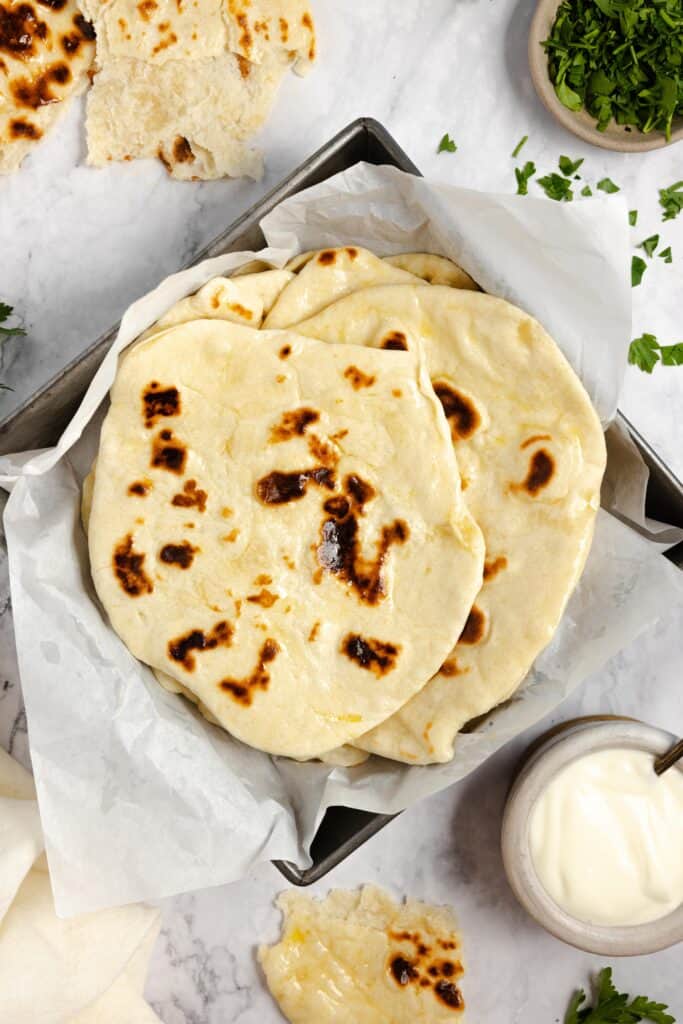sourdough naan piled on top of each other in a square baking dish lined with parchment paper on a white countertop surrounded by other ingredients