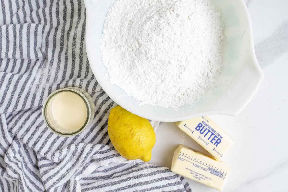 ingredients for lemon frosting, including a bowl of powdered sugar with a white and black striped tea towel with lemon, heavy cream, and butter