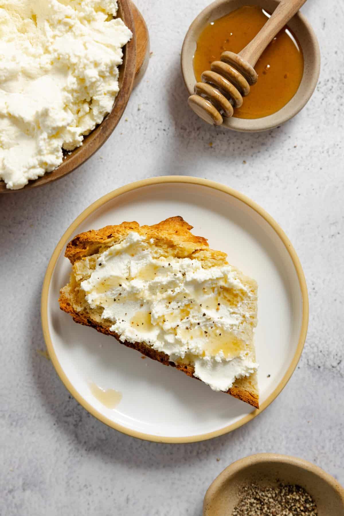 ricotta cheese slathered on toast and topped with honey on a white plate with honey and more ricotta in the background.