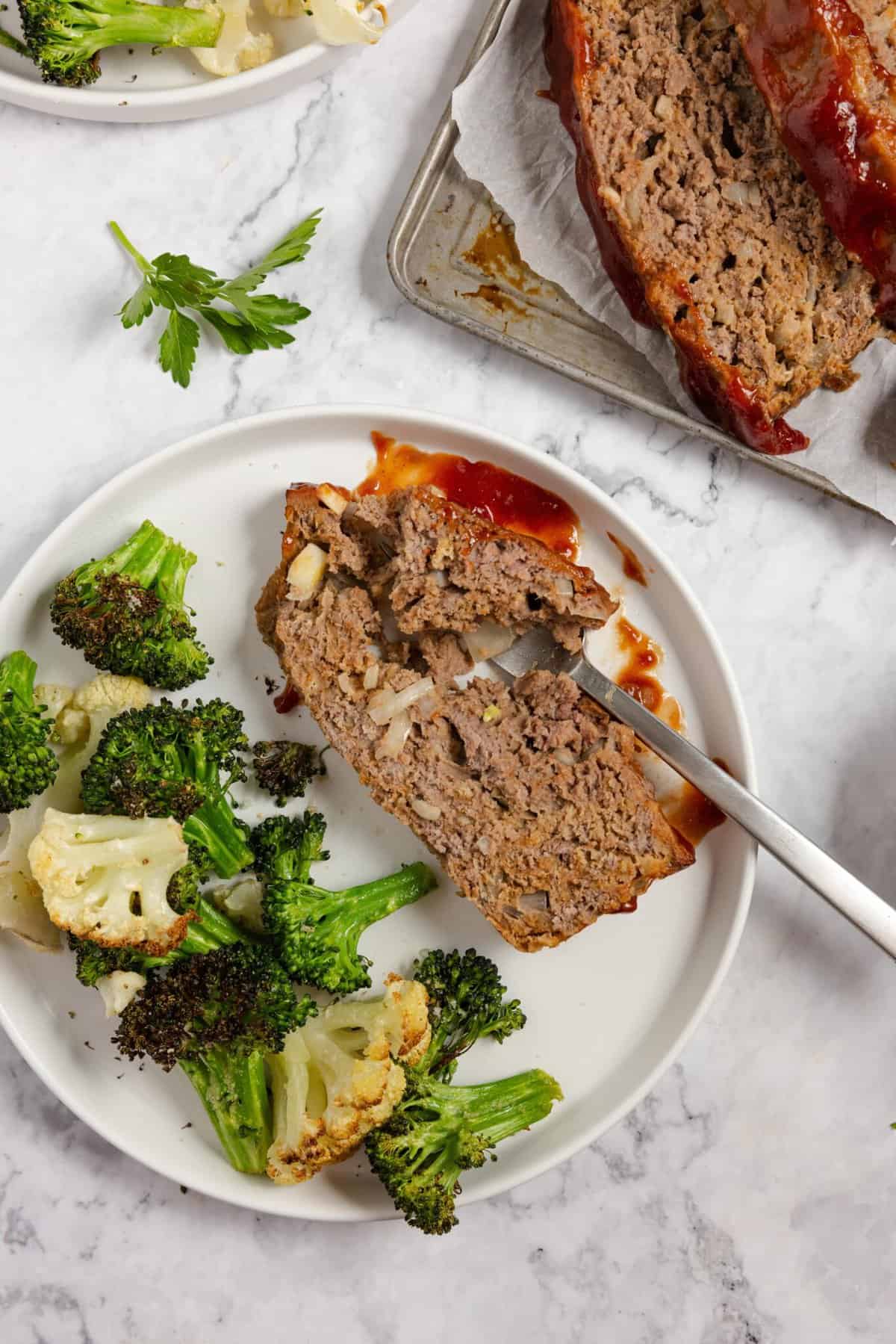 A plate with a slice of meatloaf with a fork slicing into it with a side of broccoli and cauliflower
