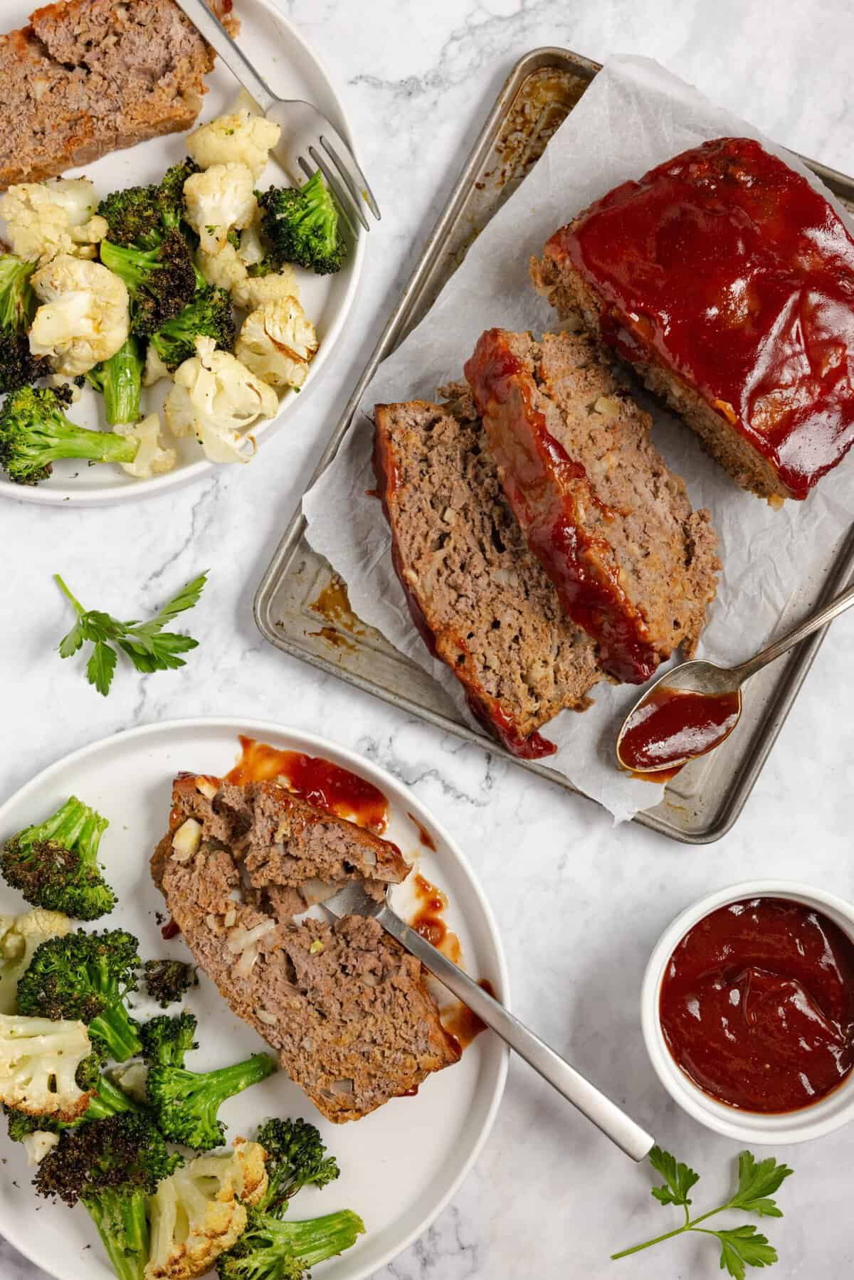 A meatloaf sliced on a plate with one slice on a separate plate next to a side of veggies on a white countertop