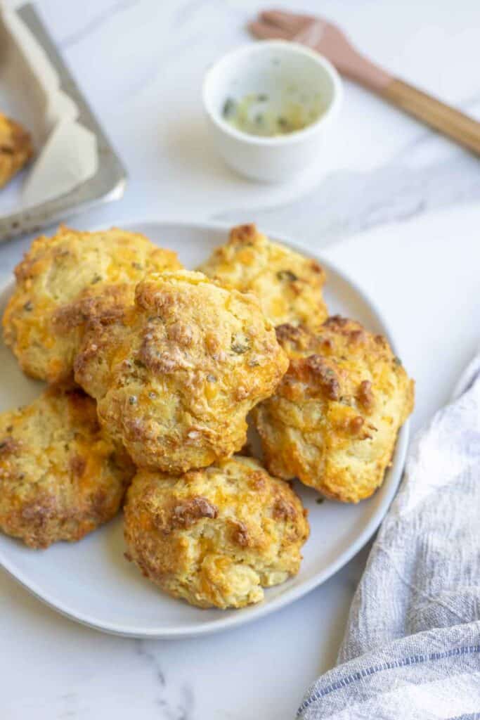 A white plate full of sourdough cheddar biscuits on a white countertop next to a tea towel and white bowl full of melted buttery and a pastry brush