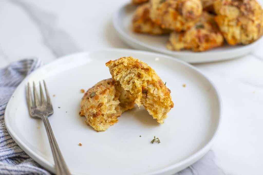 a sourdough cheddar biscuit split in half on a white plate with a silver fork and a plate full of biscuits in the background on a white countertop