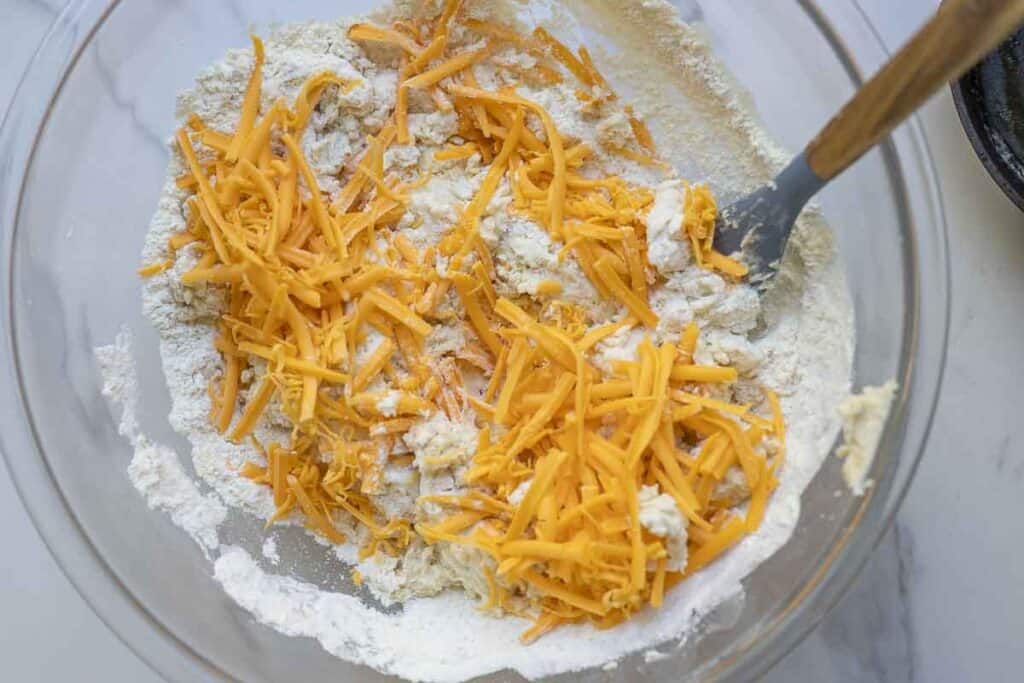 Shredded cheese added to a large glass mixing bowl full of sourdough cheddar biscuit dough ingredients with spatula 