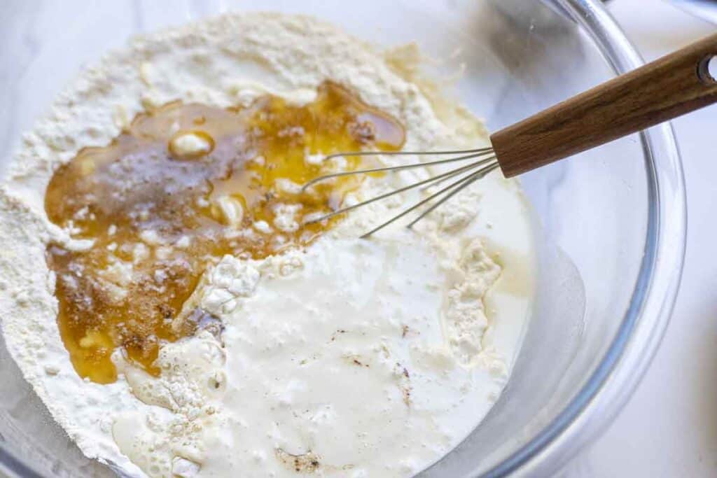 The wet ingredients added to the dry ingredients in a large glass mixing bowl with a whisk in it