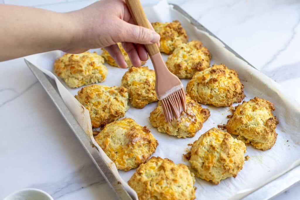 A hand brushing melted herb butter on sourdough cheddar biscuits on a baking sheet