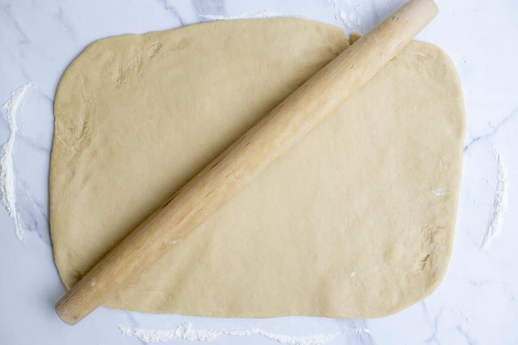 dough rolled out into a large rectangle on a marble countertop with a rolling pin on top